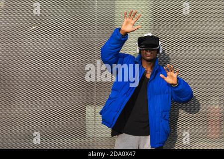 African American male standing on street near metal fence while exploring virtual reality in innovative goggles and touching air Stock Photo