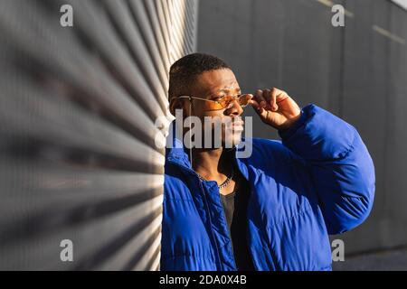 Confident African American male wearing trendy orange sunglasses and warm jacket leaning on building in city and looking away Stock Photo