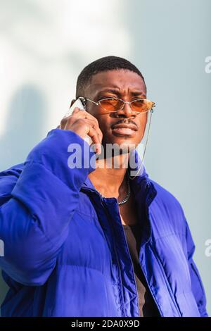 Cool handsome African American male in warm blue jacket standing on street speaking on the mobile phone looking away on sunny day Stock Photo