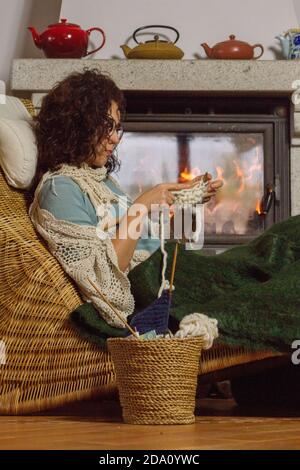 Female with glasses knitting by the chimney sat on a rattan chair. Stock Photo