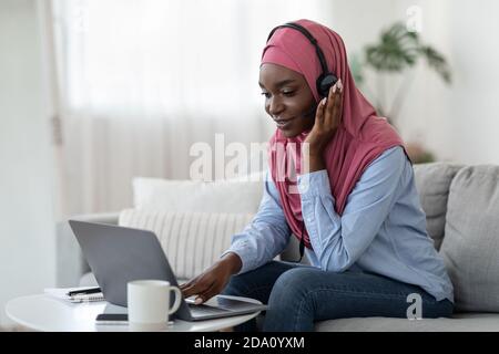 Black Muslim Female In Hijab Studying With Laptop At Home, Using Headset Stock Photo