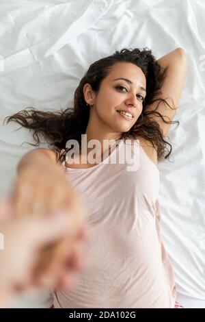 From above of crop man holding hand of charming Woman lying on soft bed at home and looking at camera Stock Photo