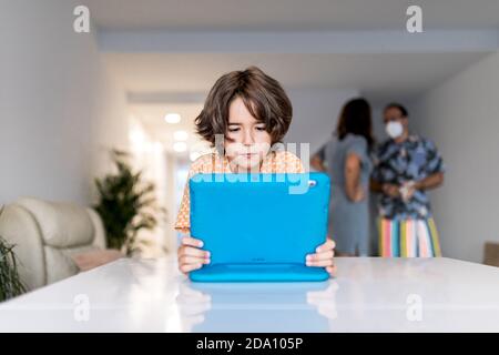 Concentrated child with netbook at table behind anonymous mother and father in mask in house during COVID 19 pandemic Stock Photo
