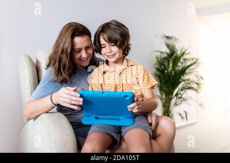 Cheerful kid with bright netbook sitting on couch with content mother in house Stock Photo
