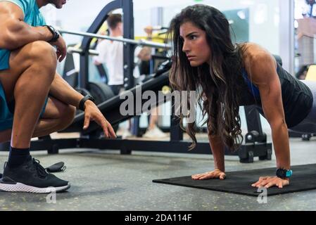 Focused ethnic female athlete doing push ups on mat under control of crop unrecognizable personal trainer in modern gym Stock Photo