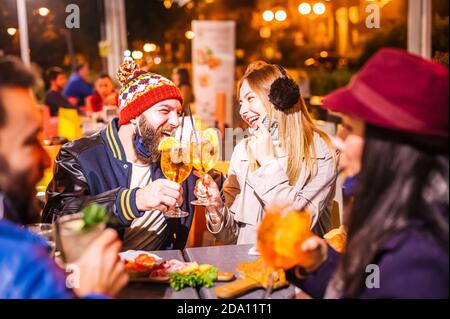Man and woman smiling and toasting spritz at winter cocktail bar outdoors wearing open face mask - New normal lifestyle concept with millennial people