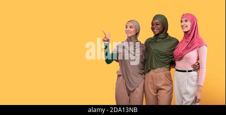 Three Muslim Ladies Pointing Finger Aside Over Yellow Background, Panorama Stock Photo