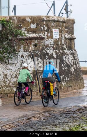 father and son cycling along the sea front past the royal yacht squadron at cowes on the isle of wight on a rainy wet day Stock Photo
