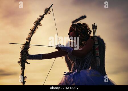 Silhouette of a cupid. Side view of teen girl archer against sunset. Little Cupid girl aiming at someone with an arrow of love. Stock Photo