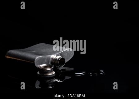 Small worn metallic hip flask spilled over on a dark reflective surface. Drinking metalic liquid isolated alcohol flask Stock Photo