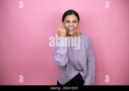 Beautiful woman wearing a casual violet sweater over pink background angry and mad raising fist frustrated and furious while shouting with anger. Rage Stock Photo