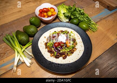 A plate of delicious smoky vegan chilli in a wrap and the ingredients used to make it on a wooden kitchen work top Stock Photo