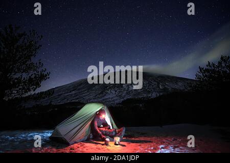 man resting in his illuminated tent on the snow under starry sky, on background Etna Mount, Sicily Stock Photo