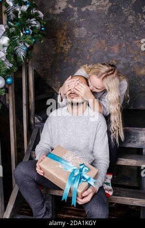 Young woman covering man's eyes with hand and surprising a man with a Christmas present. Stock Photo