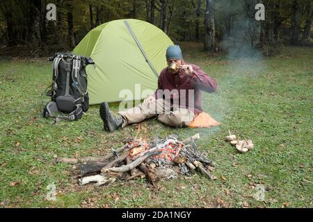 man sitting and drinking on grass of Nebrodi Park near tent, backpack and campfire, Sicily Stock Photo