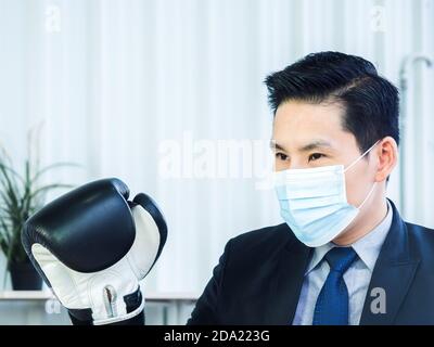 Asian Businessman wearing suit and protective face mask and hand wearing boxing gloves in office with copy space. Man wears mitten, posture in boxing Stock Photo