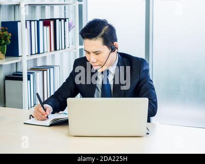 Asian businessman in suit with headset using laptop on desk in office. Man in headphones with microphone making notes and using laptop computer with c Stock Photo