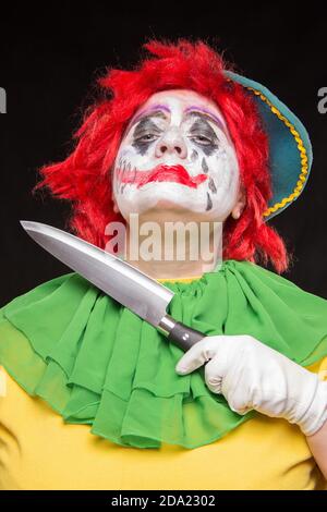 Scary clown with a horrible make-up laughs and with big knives in hands on a black background Stock Photo