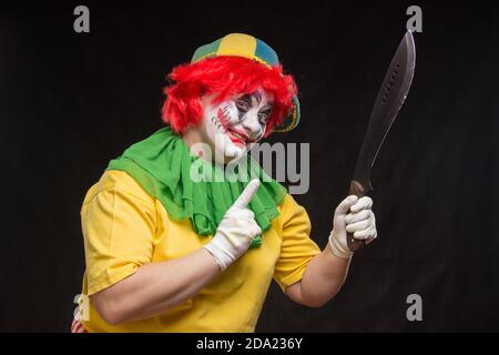 Scary clown with a horrible make-up laughs and with big knives in hands on a black background Stock Photo