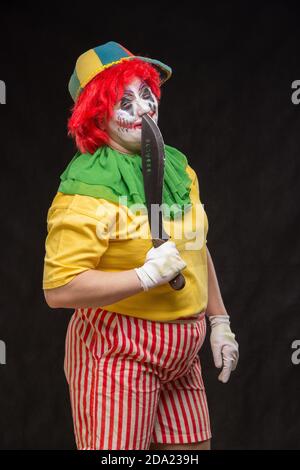 Scary clown with a horrible make-up laughs and with big knives in hands on a black background Stock Photo