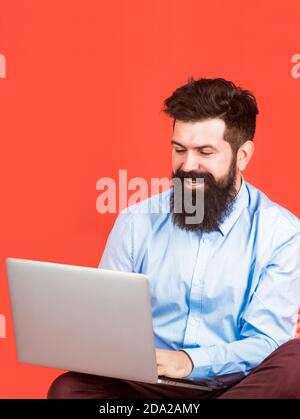 Happy young man sitting on the floor with and using laptop computer on red background. Holding laptop computer Stock Photo