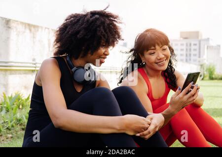 Diverse group of two female fitness friends talking and using a mobile phone in the park. Young women resting and smiling outdoors Stock Photo