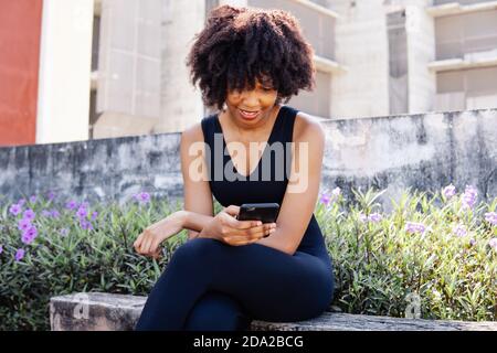 Young African American woman using a mobile phone and sitting on a bench in the park. Female black jogger resting and smiling outdoors Stock Photo