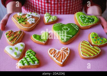 Girl holding a tray of gingerbread. In hands of women baking handmade. Painted gingerbread in form of hearts and flowers. Green and yellow cookies dec Stock Photo