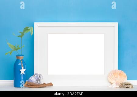 Maritime horizontal picture frame with matte mockup, fern leaves in vase, sea shells in front of blue wall. Blank image area masked with clipping path Stock Photo