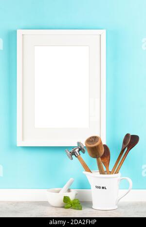 Vertical white picture frame with 2x3 aspect ratio matte on blue kitchen wall mockup . Measuring cup with cooking utensils and mortar with basil leave Stock Photo