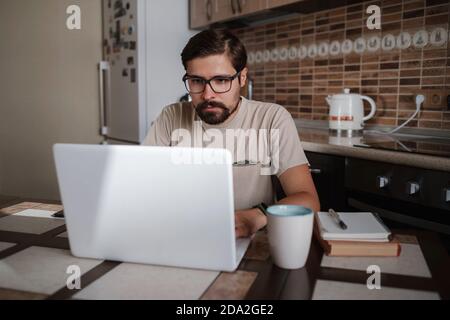 Focused young man wearing glasses using laptop, typing on keyboard, writing email or message, chatting, shopping, successful freelancer working online Stock Photo
