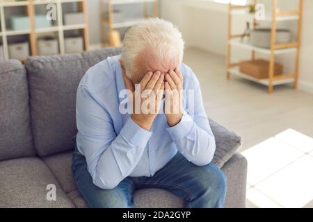 Shocked senior man covers his face with his hands from grief sitting at home on the couch. Stock Photo