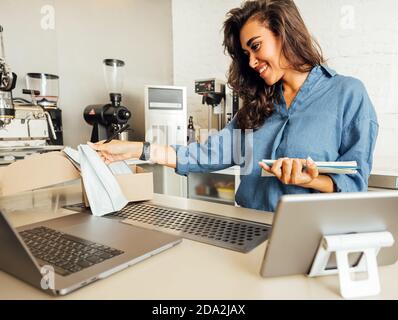 Young entrepreneur looking on pack of coffee and taking notes. Small cafe owner working at table with laptop and digital tablet. Stock Photo