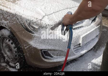 Car wash, white soap foam flies from the pump to the hood and headlight of the car, car washer at work, horizontal photography Stock Photo