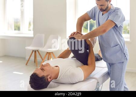 Serious doctor helping male athlete do physical exercise during rehabilitation after leg injury