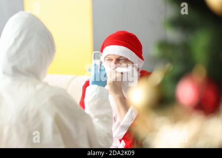 Doctor in protective suit and rubber gloves measuring temperature of sick Santa Claus with an infrared non-contact thermomete Stock Photo