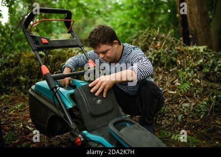 Down syndrome adult man emptying grass box in lawn mower outdoors in backyard, helping with housework concept. Stock Photo