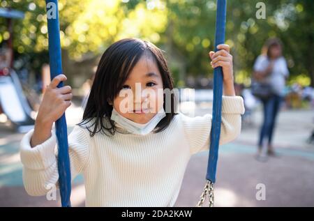 Front view portrait of small Japanese girl on swing outdoors in town, coronavirus concept. Stock Photo