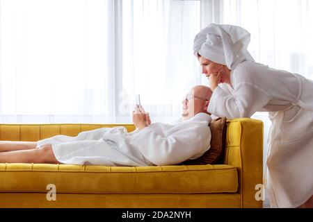 Couple in bathrobes is relaxing on the yellow sofa with mobile phone. Communication concept. Stock Photo