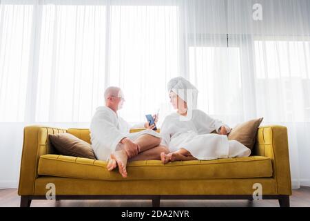 Couple in bathrobes is relaxing on the yellow sofa with mobile phone. Communication concept. Stock Photo