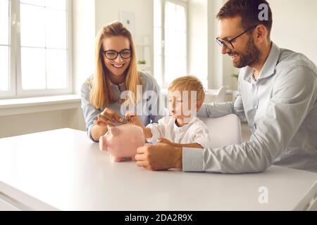 Little boy with his parents puts coins in a piggy bank sitting at a table in the room. Stock Photo