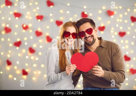Young happy loving couple in heart shaped glasses standing and holding big red heart in hands Stock Photo