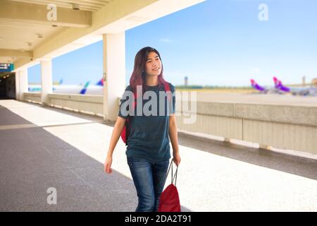 Teen girl carrying red backpack and bag walking through outdoor airport terminal with blurred airplanes in background on sunny day Stock Photo