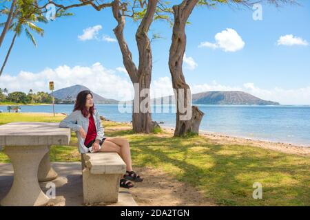 Biracial teen girl sitting on bench relaxing at shaded park next to blue ocean on island of Oahu, Hawaii with Koko Crater in background Stock Photo