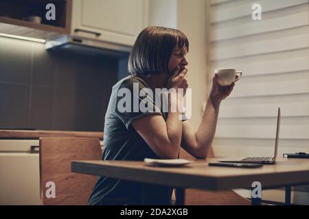 Tired exhausted female freelancer working from home, drinking coffee and yawning Stock Photo