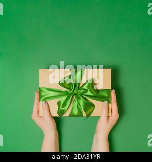 Womans hands holding craft present box with ribbon as a presenf for Christmas, new year, birthday on green background. Flat lay top view, copy space Stock Photo