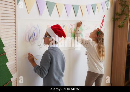 Back view portrait of boy and girl drawing on walls while wearing Santa hats and antlers for Christmas, copy space Stock Photo