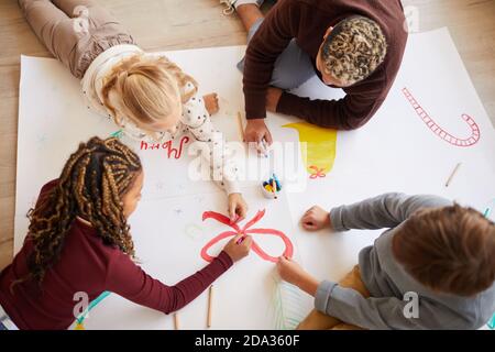 Above view portrait of female teacher sitting on floor with multi-ethnic group of kids drawing pictures while enjoying art class, copy space Stock Photo