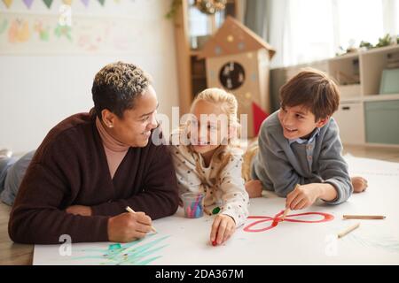 Portrait of smiling female teacher lying on floor with two kids drawing pictures while enjoying art class on Christmas, copy space Stock Photo