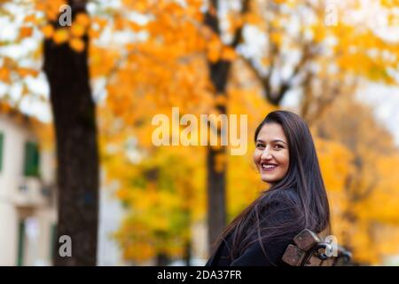 Portrait of a young woman in autumn. Scenery with warm light and falling leaves. She looks at the camera and is sitting on a bench. Stock Photo